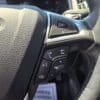 Close-up of the 2017 Ford Edge SEL steering wheel with control buttons for phone, volume, voice command, and navigation. Sunlight highlights its textured leather surface.