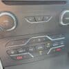 Dashboard close-up of the 2017 Ford Edge SEL showing volume and tune knobs, media controls, and a climate panel with temperature, fan, air circulation, defrost, and heated seat buttons.