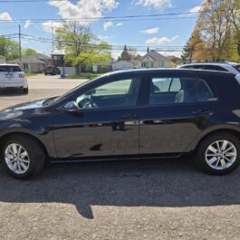 A black, four-door hatchback car is parked in a lot on a sunny day, with several other vehicles and residential houses visible in the background.