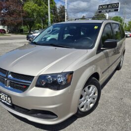 A beige 2015 Dodge Grand Caravan minivan is parked on a lot in front of a dealership called Boss Auto, under a partly cloudy sky.