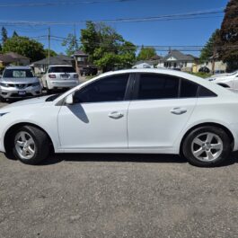 A 2016 Chevrolet Cruze 2LT Limited in white is parked on a paved lot under a clear blue sky, with other vehicles and residential houses visible in the background.