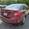 A 2012 Honda Civic LX sedan with a maroon finish is parked in a lot, displaying a Low KM sign on the license plate. Other vehicles and green trees can be seen in the sunny background.