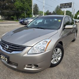 A 2012 Nissan Altima 2.5 SL is parked on a gravel lot outside Boss Auto dealership, with trees and other vehicles in the background on a sunny day.