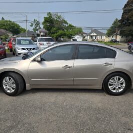 A 2012 Nissan Altima 2.5 SL in silver is parked in a lot among several other cars, with houses and trees visible nearby on a cloudy day.