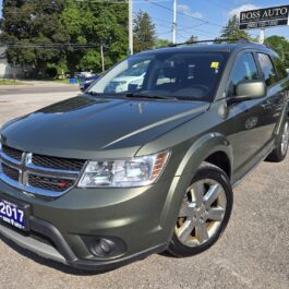 A metallic green 2017 Dodge Journey SUV is parked in a lot in front of a car dealership named Boss Auto, under a clear blue sky. The vehicles front and side are visible, with the headlights off.