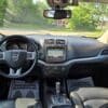 Interior of a 2017 Dodge Journey Crossroad V6 AWD showcasing the steering wheel, dashboard, navigation touchscreen, gear shift, and YOUR BUSINESS IS APPRECIATED! floor mats with sunlight highlighting its AWD capability.