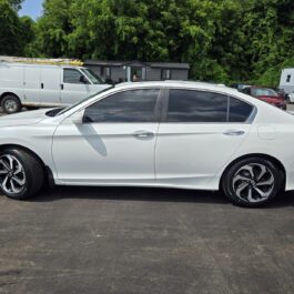 A 2016 Honda Accord EX-L white sedan with tinted windows is parked on asphalt near trees, a white van, and several vehicles, with small buildings or trailers in the background.