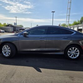 A 2015 Chrysler 200 C sedan in dark gray is parked on asphalt under a sunny sky, with buildings, trees, and other vehicles visible in the background.