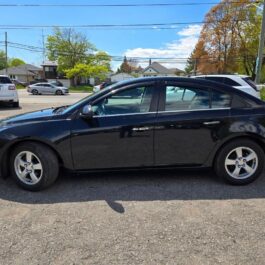 A black four-door sedan is parked in a lot on a sunny day, surrounded by other vehicles. Houses, trees with green foliage, and a clear blue sky are visible in the background.