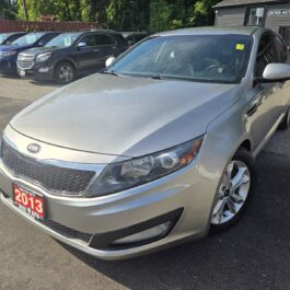 A 2013 Kia Optima EX sedan in silver is parked outside on a sunny day, with other cars behind it. A red dealer plate on the front highlights the model year 2013.