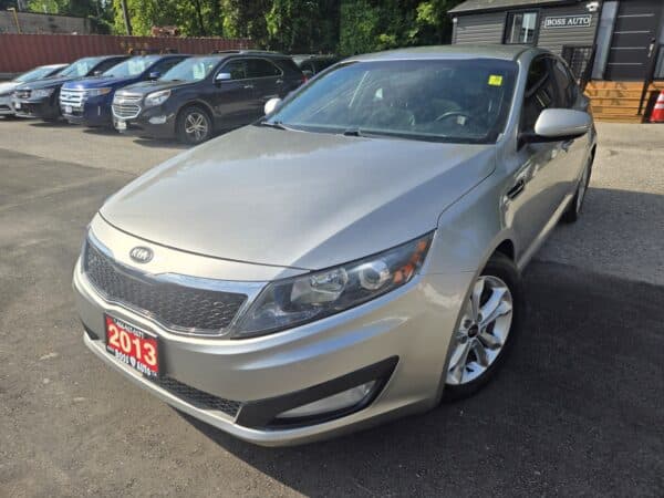 A 2013 Kia Optima EX sedan in silver is parked outside on a sunny day, with other cars behind it. A red dealer plate on the front highlights the model year 2013.