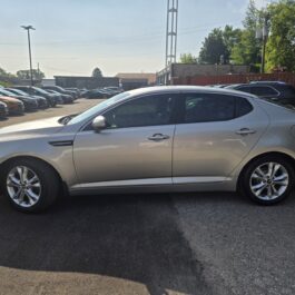 A 2013 Kia Optima EX is parked in a car lot on a sunny day, with other vehicles and trees visible in the background.