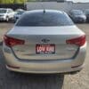 A silver 2013 Kia Optima EX with tinted windows is parked in a lot. A red and white LOW KMS sign with dealership info is on the back, and several other cars are visible in the background.