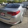A silver 2013 Kia Optima EX with a LOW KMS sign on the rear is parked among other cars under a sunny sky, with trees and a white building in the background.