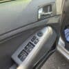 Close-up of the 2013 Kia Optima EX driver-side door panel featuring power window controls, side mirror adjustment switch, door handle, and a floor mat visible outside through the open door.