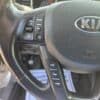 Close-up of the 2013 Kia Optima EX steering wheel features control buttons for volume, mode, eco, trip, and reset. The car door is open with parts of the dashboard and pedals seen in the background.