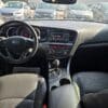 View of a 2013 Kia Optima EX interior from the back seat, featuring the steering wheel, dashboard, center console with controls and gear shift, sunlight on black leather seats, and parked cars visible through the windshield.