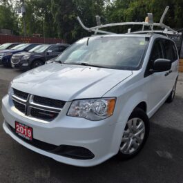 A 2019 Dodge Grand Caravan SE Plus CARGO in white with a roof rack is parked in a lot, surrounded by other vehicles and trees.