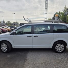 A white 2019 Dodge Grand Caravan SE Plus CARGO with roof cargo equipment and a yellow beacon is parked in a lot among other vehicles under a cloudy sky.