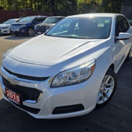 A 2015 Chevrolet Malibu LT sedan with a white exterior is parked in a sunny lot among other cars, showing its front and driver’s side. A red license plate highlights the model year 2015.