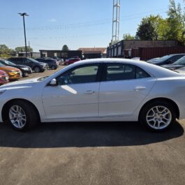 A 2015 Chevrolet Malibu LT is parked in a car lot on a sunny day, with other vehicles nearby and trees and buildings visible in the background.