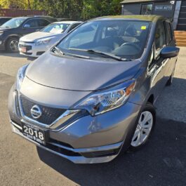 A gray 2018 Nissan Note hatchback is parked in a lot with other cars nearby, under bright sunlight. The car has a dealership license plate and is displayed outdoors near a building.