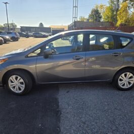 A gray compact hatchback car is parked in a lot on a sunny day, surrounded by other vehicles and trees in the background.