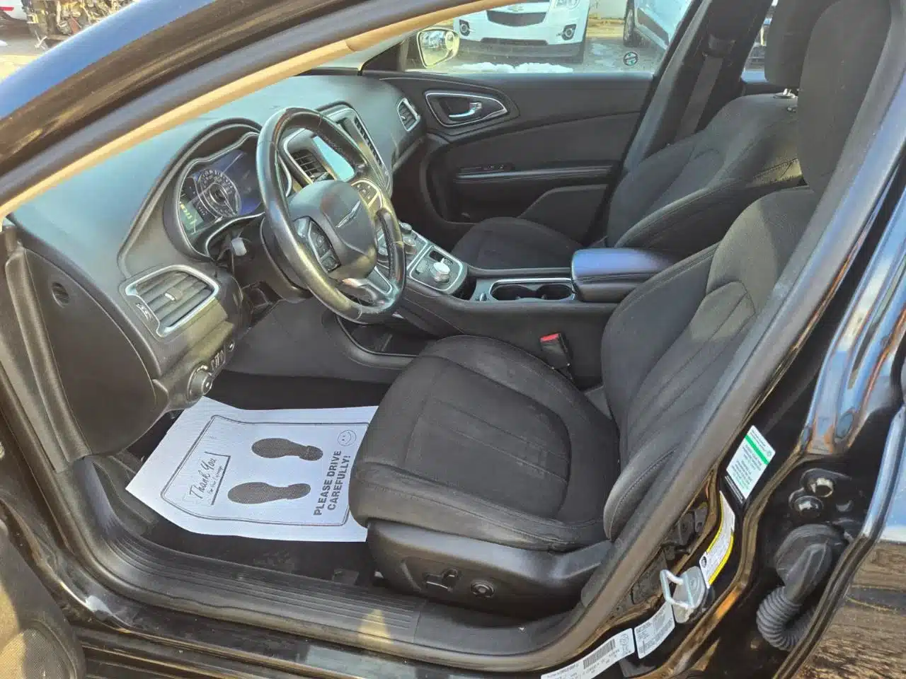 Inside the 2015 Chrysler 200 Limited, the driver’s seat, passenger seat, dashboard, steering wheel, and center console are visible. A paper floor mat with footprints is placed on the driver’s side floor.