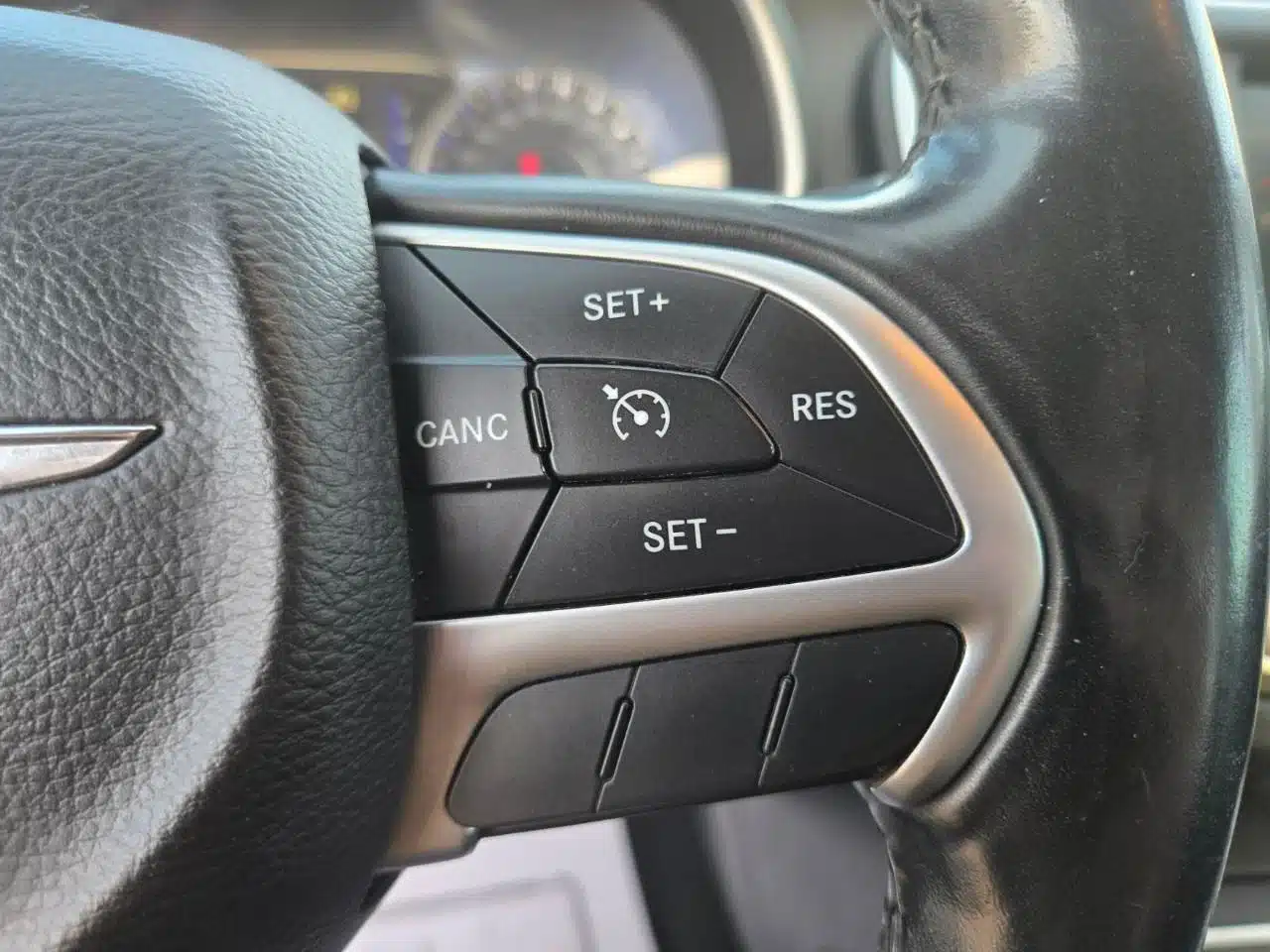 Close-up of the 2015 Chrysler 200 Limited steering wheel, displaying cruise control buttons labeled SET+, SET-, CANCEL, and RES with a speedometer icon in the center. The black leather steering wheel is accented by silver trim, showcasing the premium interior design of the 2015 Chrysler 200 Limited.