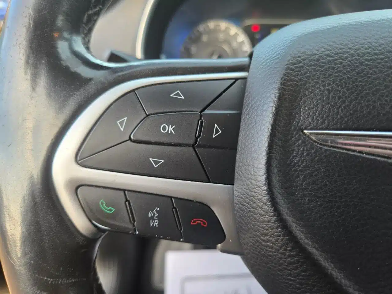 Close-up of the 2015 Chrysler 200 Limited steering wheel displaying control buttons such as directional arrows, an OK button, phone call controls, and a voice command button, with part of the dashboard visible in the background.