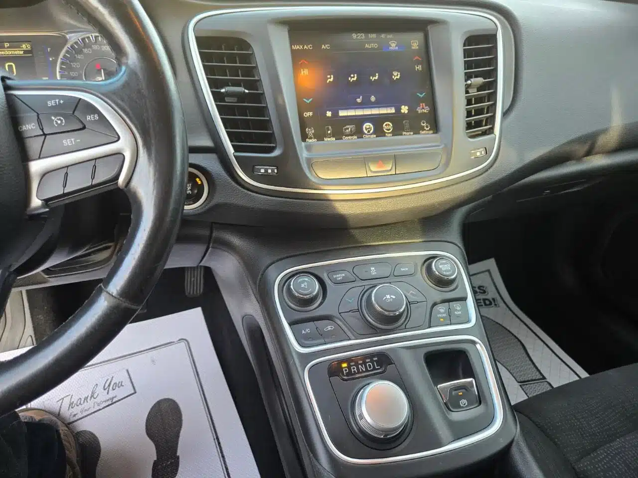 Close-up of the 2015 Chrysler 200 Limited’s center console featuring a touchscreen display with integrated climate controls, a rotary gear shift dial, drive mode buttons, and partial views of the steering wheel and dashboard. Floor mats appear below—illustrating the refined interior details of this used 2015 Chrysler 200 Limited.