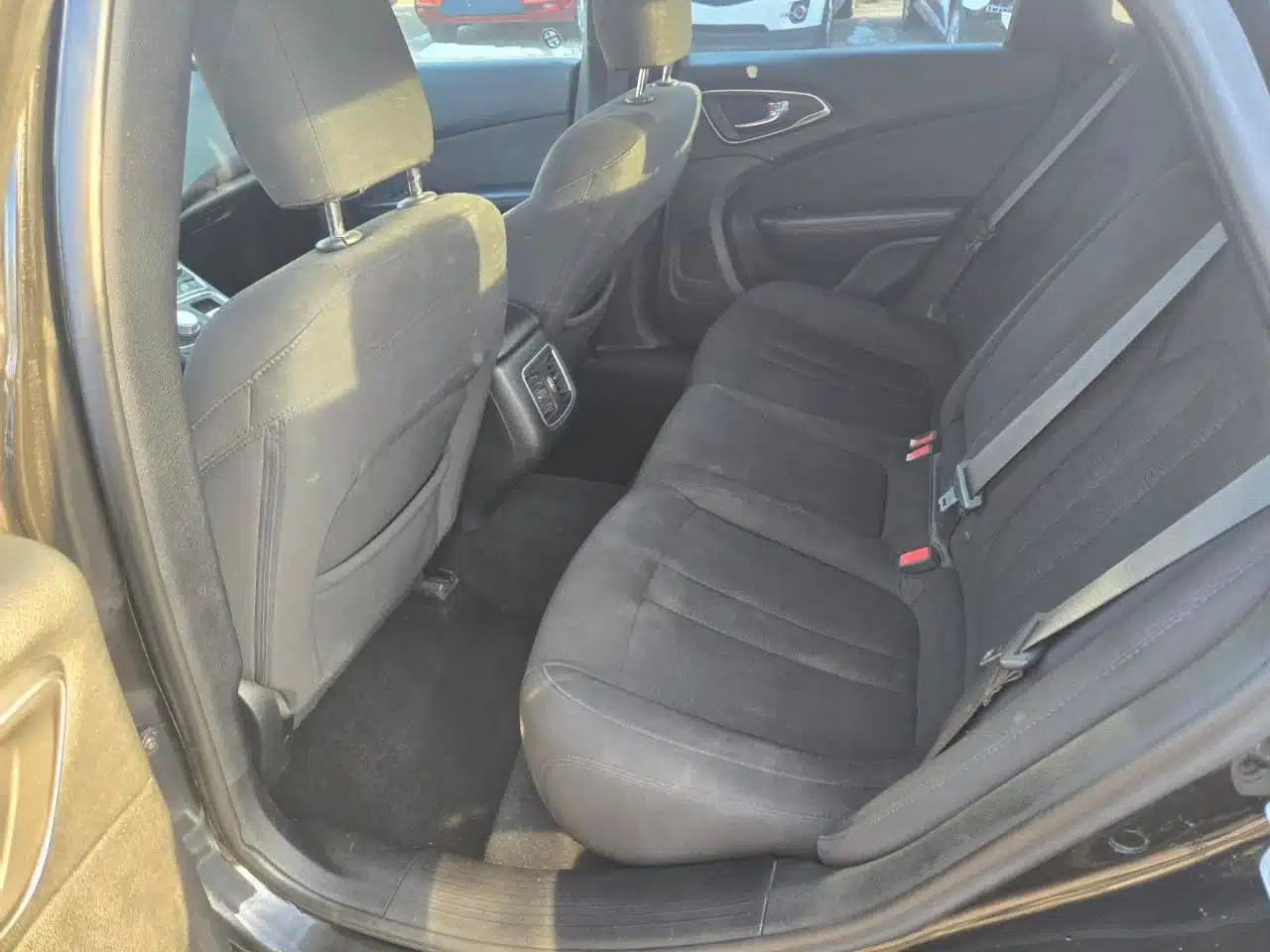 The rear seat area of this 2015 Chrysler 200 Limited features black fabric seats, two seat belts, headrests, and rear air vents, with sunlight streaming in through the windows.