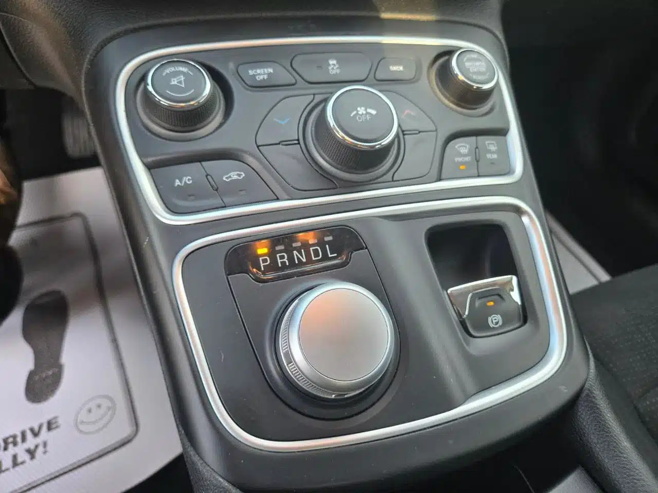 The image displays the center console of a 2015 Chrysler 200 Limited, highlighting intuitive climate controls, a gear shift dial set to P, an electronic parking brake, and multiple buttons for air conditioning, defrost, and driving modes. A partially visible floor mat can also be seen.
