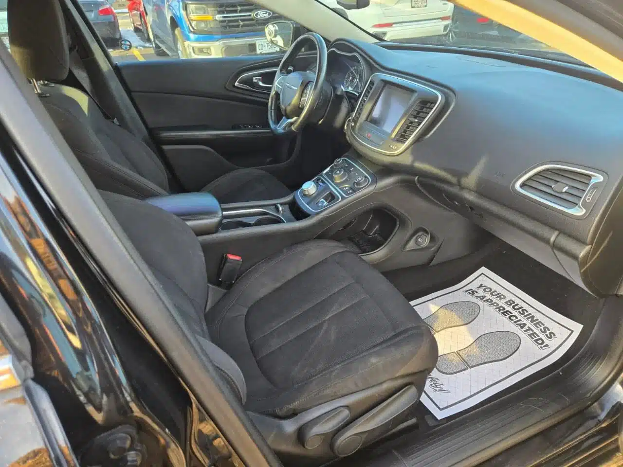 The interior of the 2015 Chrysler 200 Limited features black seats, a center console, and a dashboard display. On the passenger side floor, there is a paper floor mat with the message “Your business is appreciated!”.