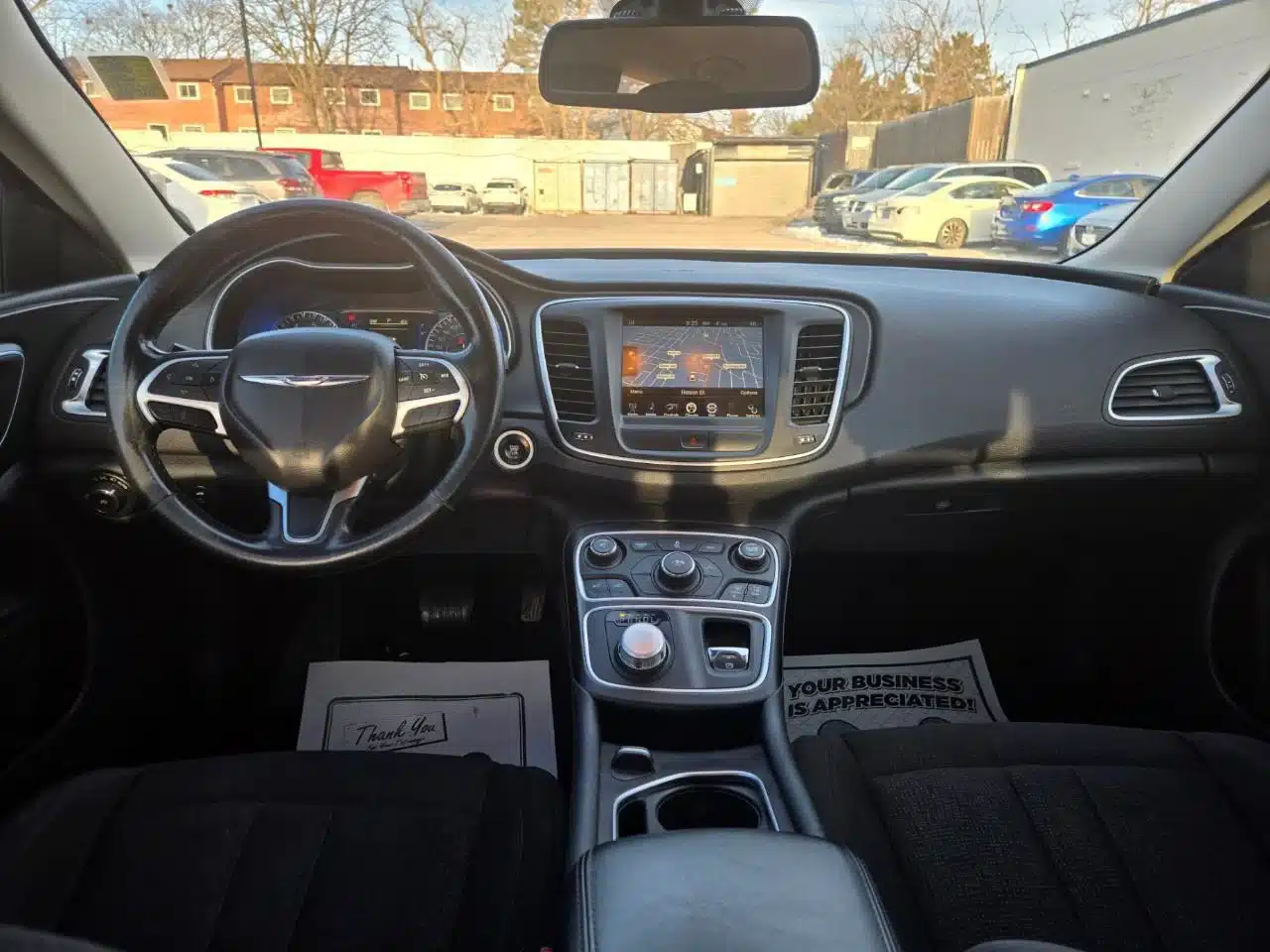 Interior view from the rear seat of a 2015 Chrysler 200 Limited, highlighting the steering wheel, dashboard with touchscreen display, gear knob, and both front seats illuminated by sunlight streaming through the windshield.