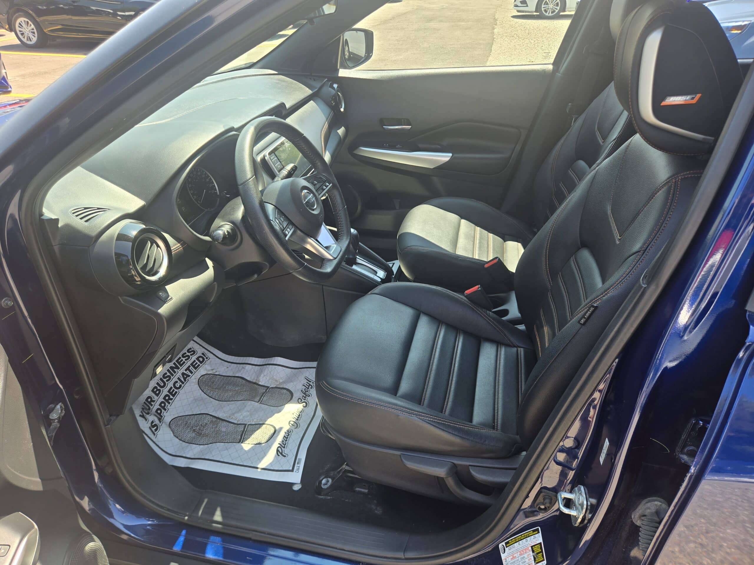 View of the 2019 Nissan Kicks SR’s black interior from the open driver’s side door, displaying the seats, dashboard, steering wheel, and a floor mat featuring “Your Business is Appreciated” with printed footprints.