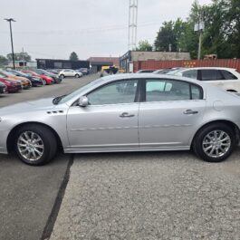 A silver sedan is parked in a lot with several other vehicles. Trees, a building, and a shipping container are visible in the background. The weather appears to be overcast.