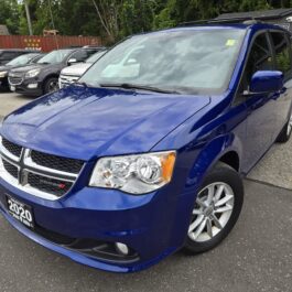A blue 2020 Dodge Grand Caravan minivan is parked in a lot with other vehicles in the background, under a cloudy sky.