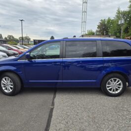 A blue minivan is parked on a lot, with other cars and trees in the background under a cloudy sky.