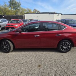 Side profile of a red sedan parked in a car lot with other vehicles in the background.
