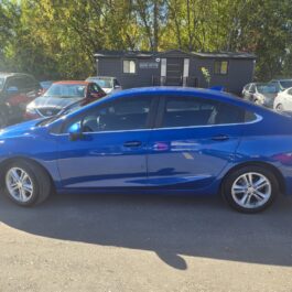 A bright blue Chevrolet Cruze sedan parked in a car lot in front of a small office building.
